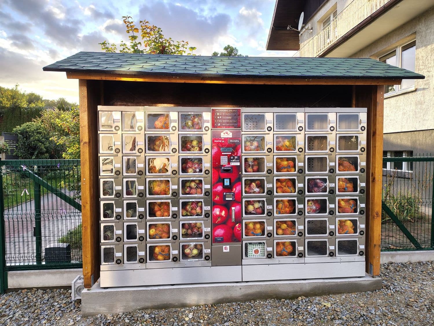 Jabłkomat Sadów Celmerów — automat ze skrytkami pełnymi jabłek pod drewnianym daszkiem przy bramie gospodarstwa